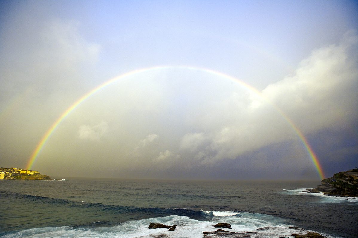 A full rainbow arching over the ocean at Bondi with headlands on either side