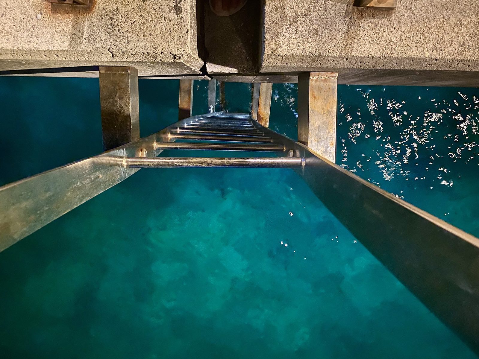 Looking down the metal ladder on the wharf at Flying Fish Cove into clear deep turquoise water, an Earth to Sea News original