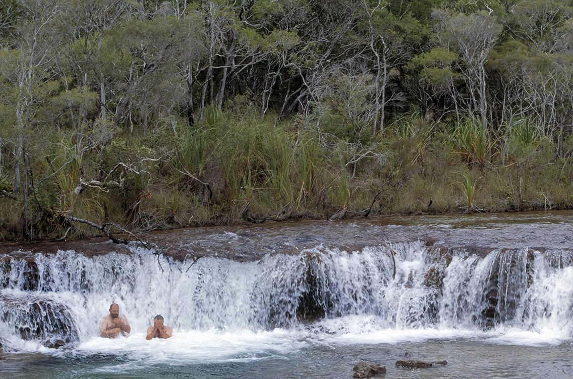 Two swimmers in the pool below a wide bush waterfall on Cape York